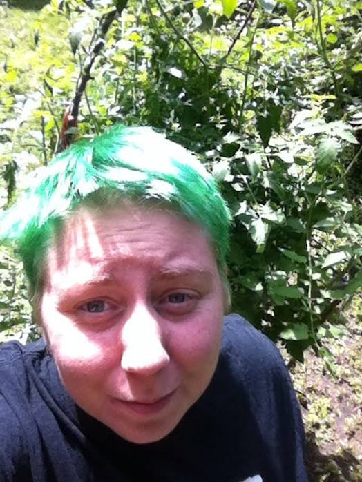 A person with green hair, and a black shirt taking a selfie in a garden