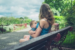 A woman in a blue top breastfeeding her baby on a park bench on a bright sunny day