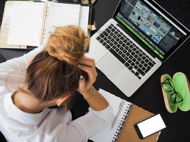 Woman holding her head in frustration while working on her laptop from home with notebooks next to h...