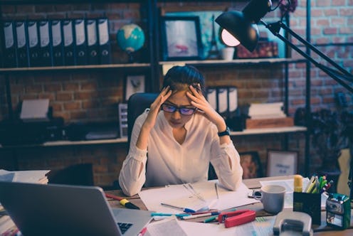 A woman holding her head over a pile of papers and a laptop on her desk