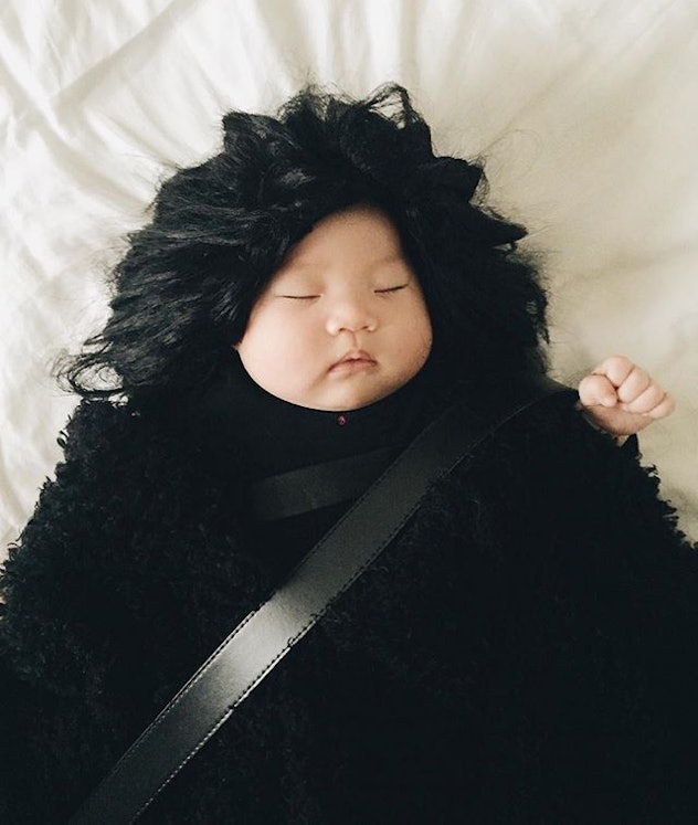A baby in a black outfit wearing the black wig sleeping on the bed