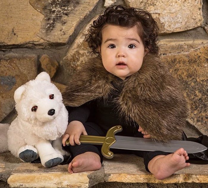 A baby dressed like Jon Snow holding a sword and sitting next to a white teddy bear