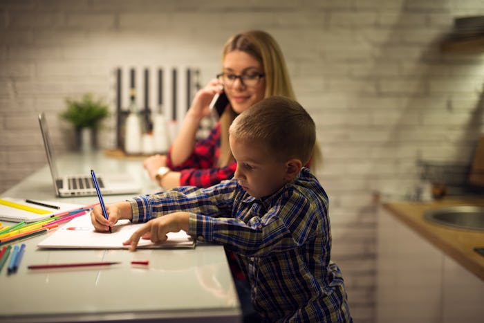 A mom working on her phone and laptop in the kitchen with her son coloring at the kitchen isle