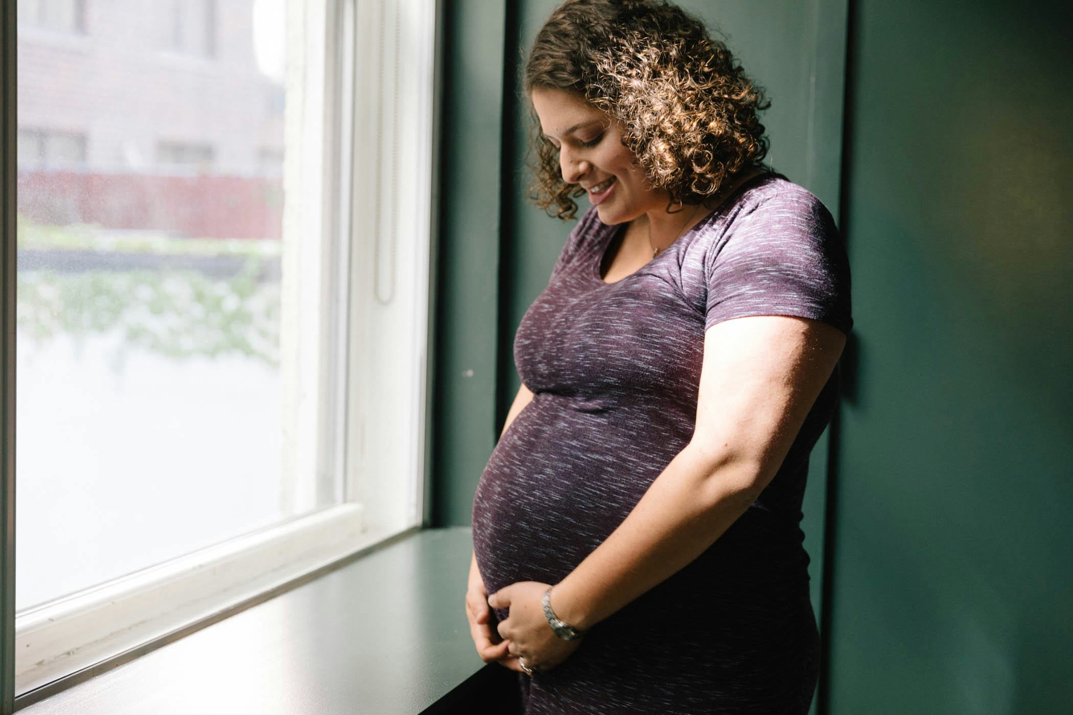 A pregnant woman feeling her baby's movements, smiling beside a window.