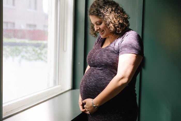 A pregnant woman feeling her baby's movements, smiling beside a window.