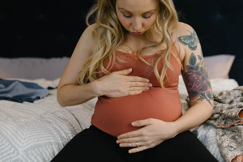 A pregnant woman with tattoos on her arm sits on the bed and holds her belly
