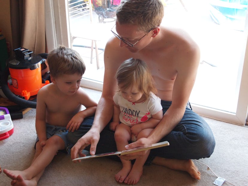 Gemma’s husband sits on the floor with their daughter in his lap and his son beside him while readin...