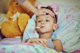 Mom putting a hand on her sons forehead to gauge his temperature while he lays in bed with a thermom...