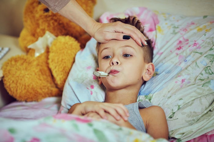 Mom putting a hand on her sons forehead to gauge his temperature while he lays in bed with a thermom...