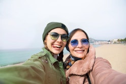 A trendy mom and daughter wearing coats and sunglasses snap a selfie on a beach.