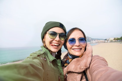A trendy mom and daughter wearing coats and sunglasses snap a selfie on a beach.