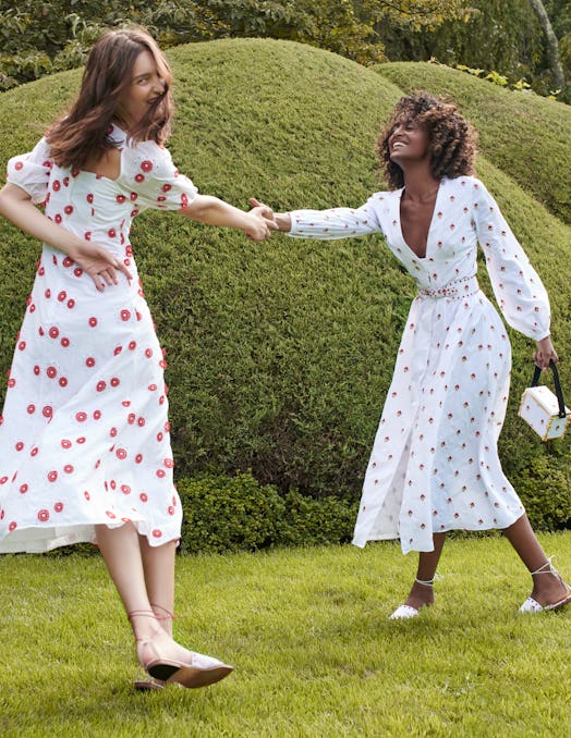 Two women wearing white dresses with flower details while holding hands in the garden.