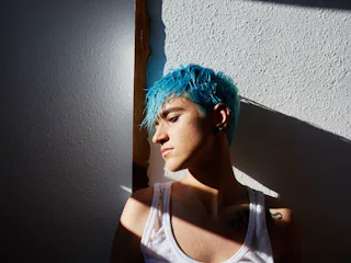A man with a blue pixie haircut standing against a wall posing for a picture.