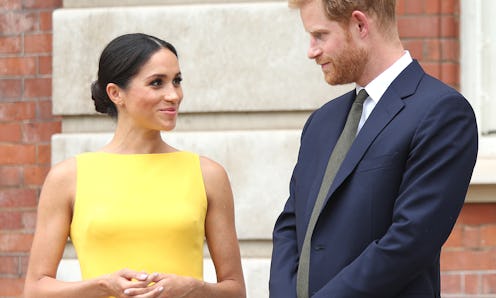 Meghan Markle smiling in a yellow dress while looking at Prince Harry in a navy suit, white shirt, a...