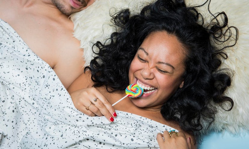 A woman biting into a lollipop while lying in bed next to a man