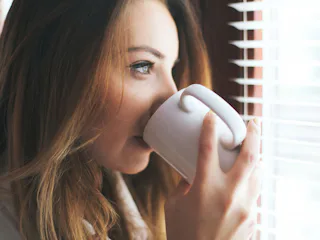 A woman drinking coffee, looking at the sunlight from her window as it has health benefits