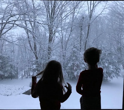 Two kids standing next to a window and looking at the snow outside during Holiday season