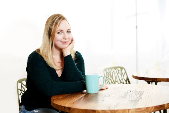 April Daniels Hussar smiling and sitting on a chair next to a wooden table with a blue mug on it