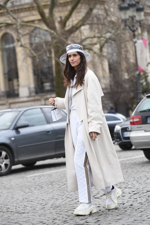 A woman wearing an off-white silky coat over white pants and top, and a clear PVC hat