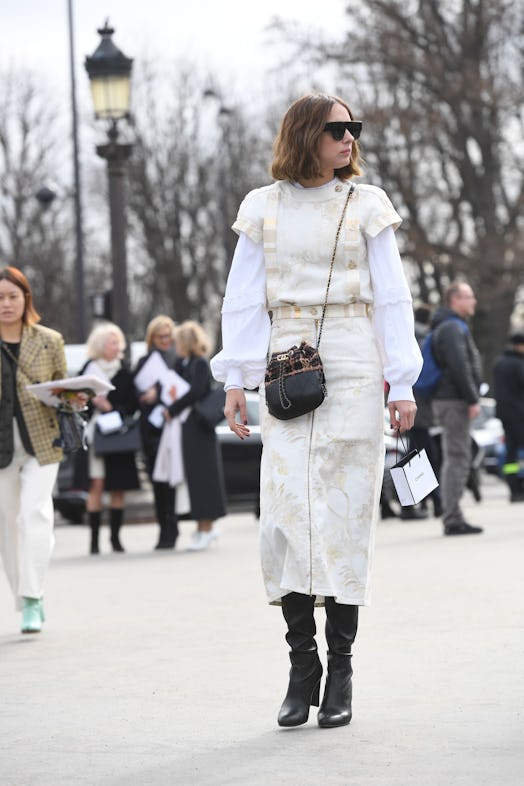 A woman wearing an off-white dress over a white silk blouse and black tall boots