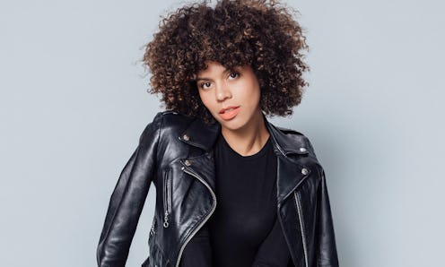 Grasie Mercedes posing with her brown curly hair in a black leather jacket in front of the grey back...