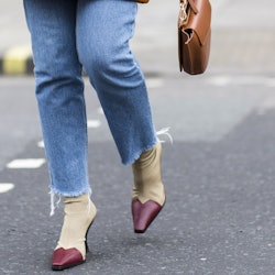 A woman crossing the street in jeans and beige and maroon ankle boots