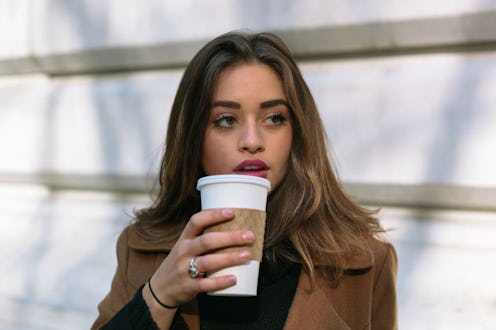A young brunette holding a large coffee-to-go, sitting outside.