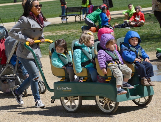 A mom pushing a green kinder van with six toddlers in it in a park