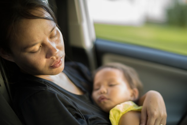 A fatigued mom with her eyes closed sits in the car holding her baby in her arms
