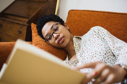 A woman on an orange couch reads a book for self-care, feeling sad that she's missing thanksgiving w...