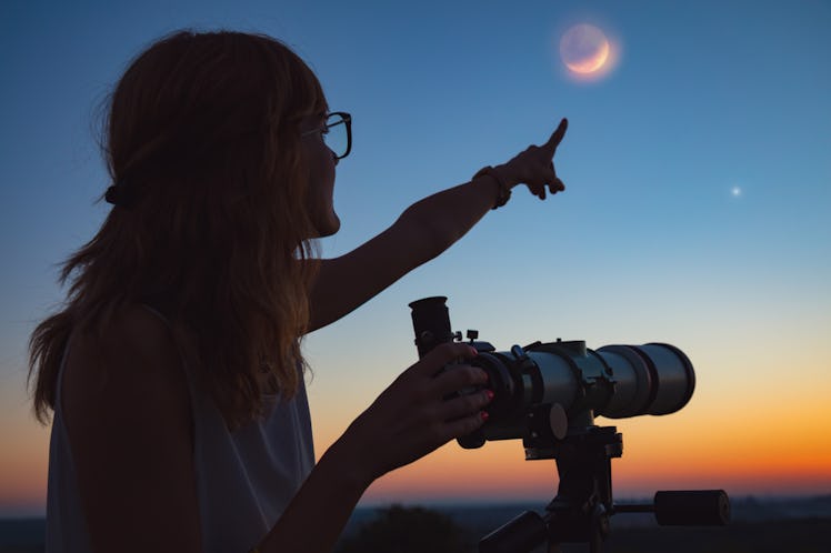 A woman points to the sky, while stargazing with a telescope at sunset.
