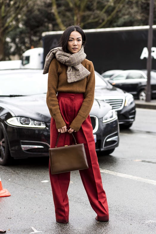 A woman standing on the street in burgundy wide leg trousers, a brown sweater and a plaid padded sca...