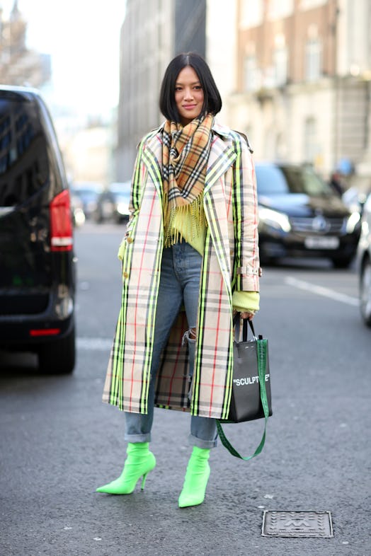A woman wearing an electric green trench coat paired with Burberry plaid scarf, neon ankle boots and...