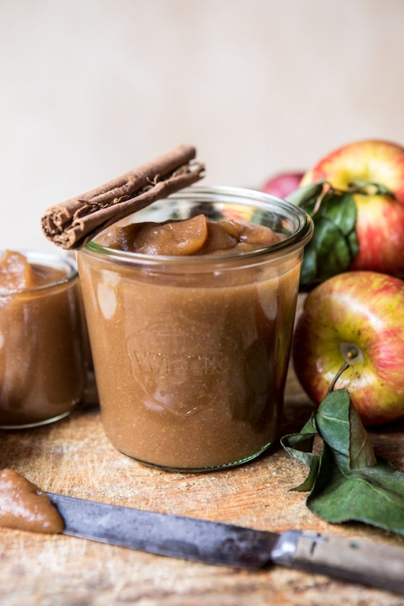 caramel-colored apple butter in a glass cup with cinnamon stick on top presented next to some apples...