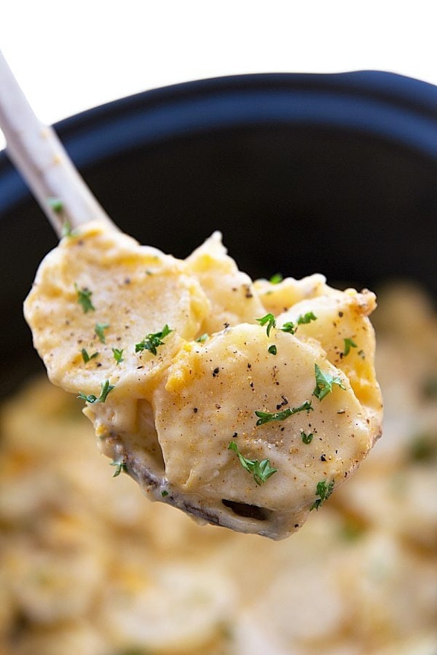 close up of creamy chunks of potatoes with seasoning and green garnish being scooped from crock pot