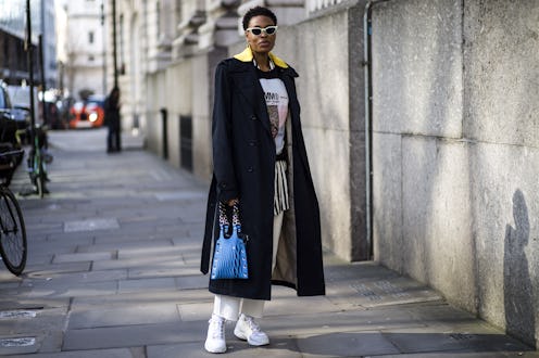 A woman wears white sneakers black coat layered over a hoodie with a print, and a stripy shirt