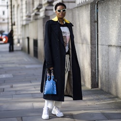 A woman wears white sneakers black coat layered over a hoodie with a print, and a stripy shirt