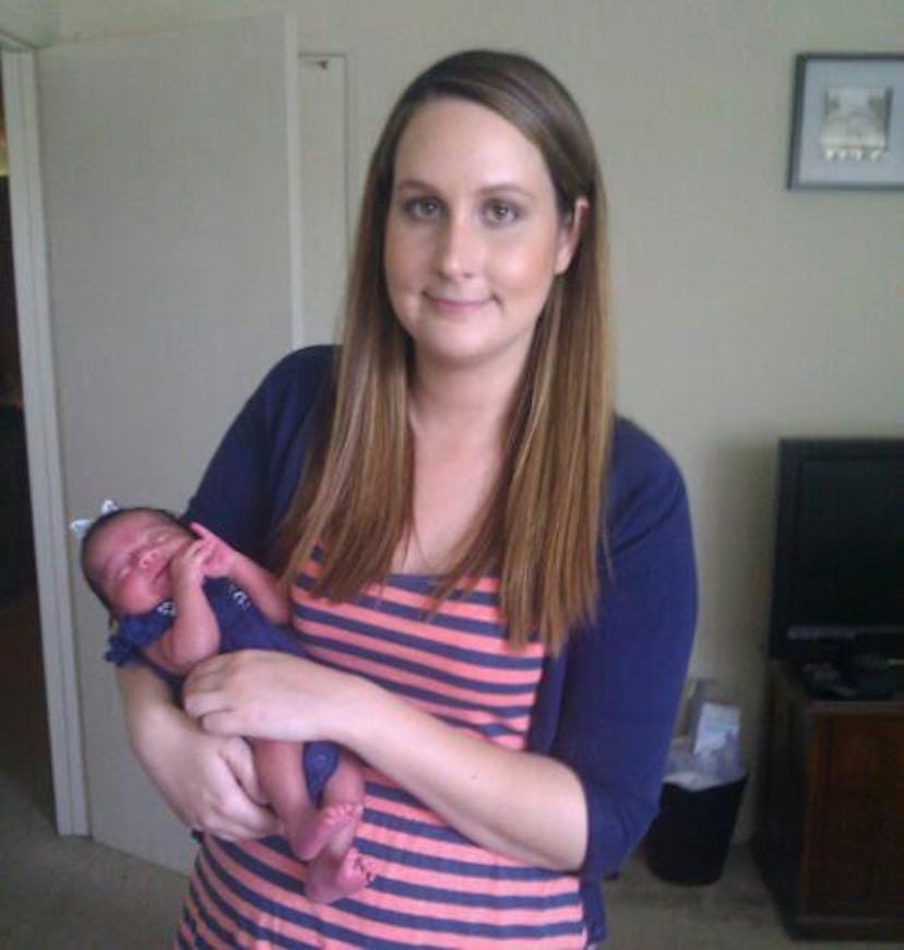 A woman with long brown hair wearing a striped shirt holding a newborn in a blue onesie.