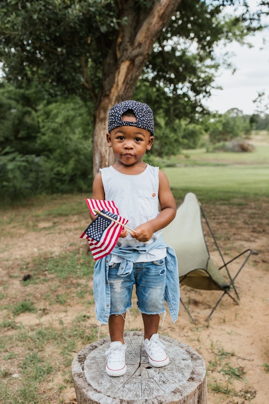A kid standing outside holding two small american flags while wearing jean shorts and a cap