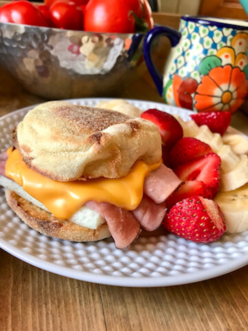english muffin with egg, ham and cheese next to sliced strawberries and bananas on a white plate