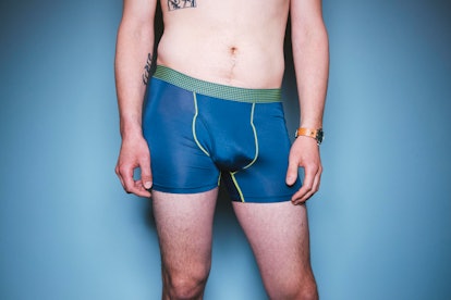 A tattooed white man poses in blue underwear, shot from his bellybutton to knees. 