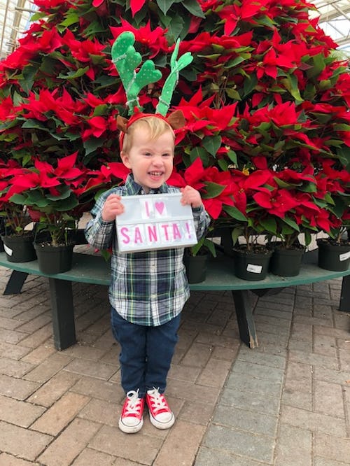 A toddler holding a sign that says I love santa in front of potted flowers stacked to look like a ch...