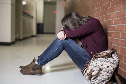 Girl sitting sad and hiding her face in the hallway, leaning against a brick wall