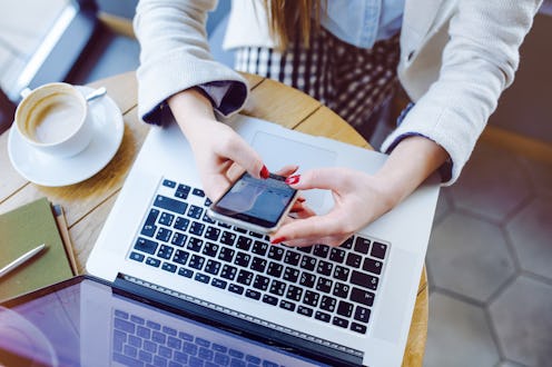 A woman typing on a phone over her laptop's keyboard in a coffee shop