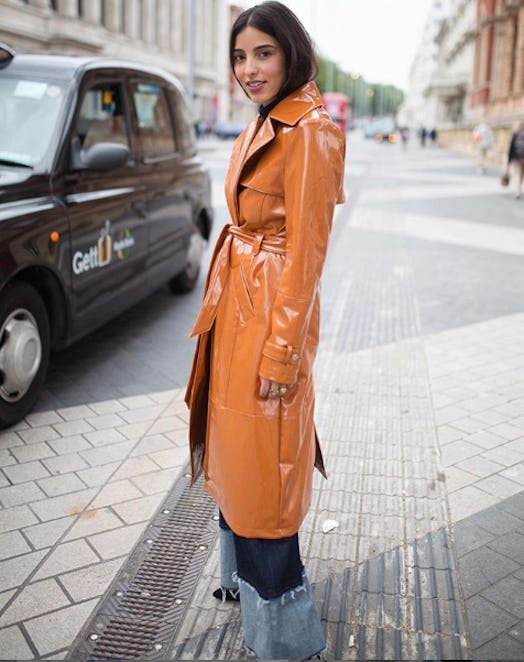 A woman in a long bronze-orange shiny leather trench coat and blue denim jeans