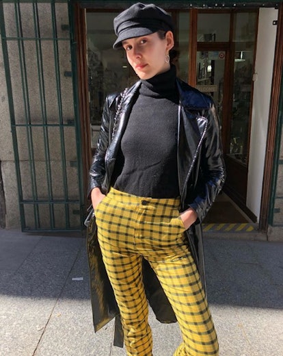 A woman in a black leather trench coat, black turtleneck, black marine cap and yellow-black checked ...