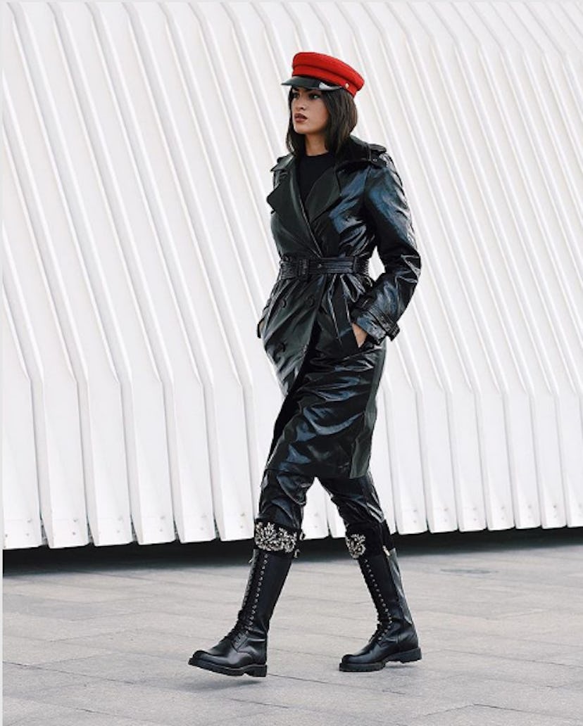 A woman in a long black leather trench coat and trousers, black boots, and a red marine cap
