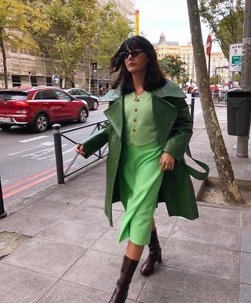 A woman in a dark green leather trench coat, light green waistcoat, burgundy boots and black sunglas...