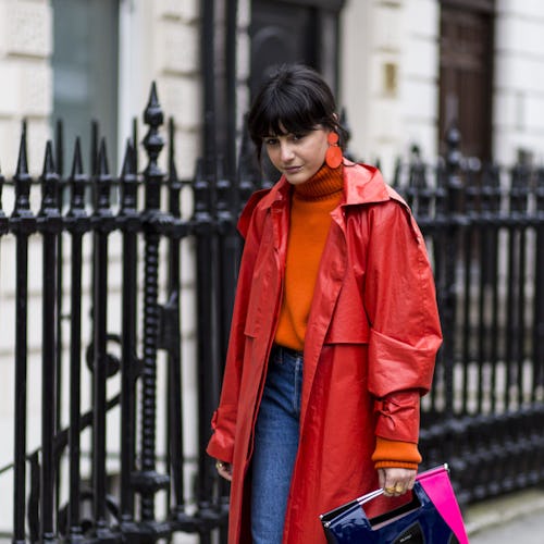A woman in a red leather trench coat, orange sweater, blue jeans, purple shoes, and a black-pink-ora...