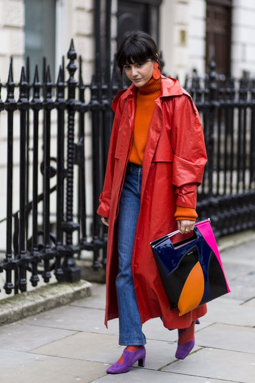 A woman in a red leather trench coat, orange sweater, blue jeans, purple shoes, and a black-pink-ora...